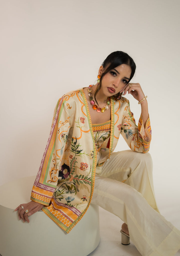 Seated front view of a pastel orange pret wear three-piece in luxury printed silk, featuring an open printed coat over a coordinating top and straight pants, from our boutique in Islamabad F-7.